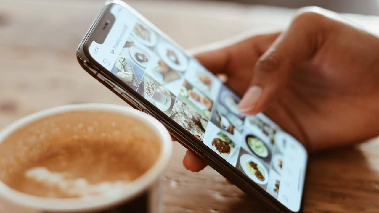 Mão segurando um smartphone, com a tela exibindo uma galeria de fotos de pratos de comida. Ao fundo, desfocado, está um copo de café.