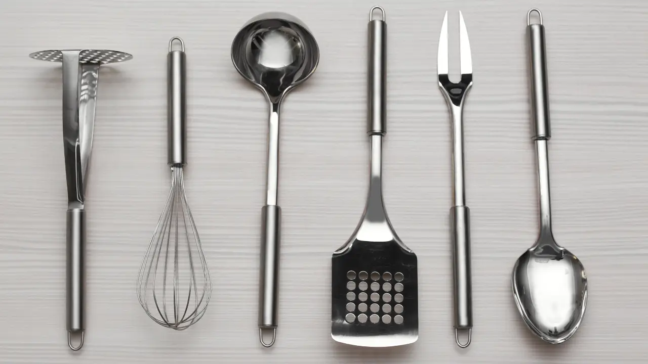 "Utensílios de cozinha de aço inoxidável organizados em uma superfície clara. Da esquerda para a direita: amassador de batatas, batedor, concha, espátula com furos, garfo trinchante e colher grande.