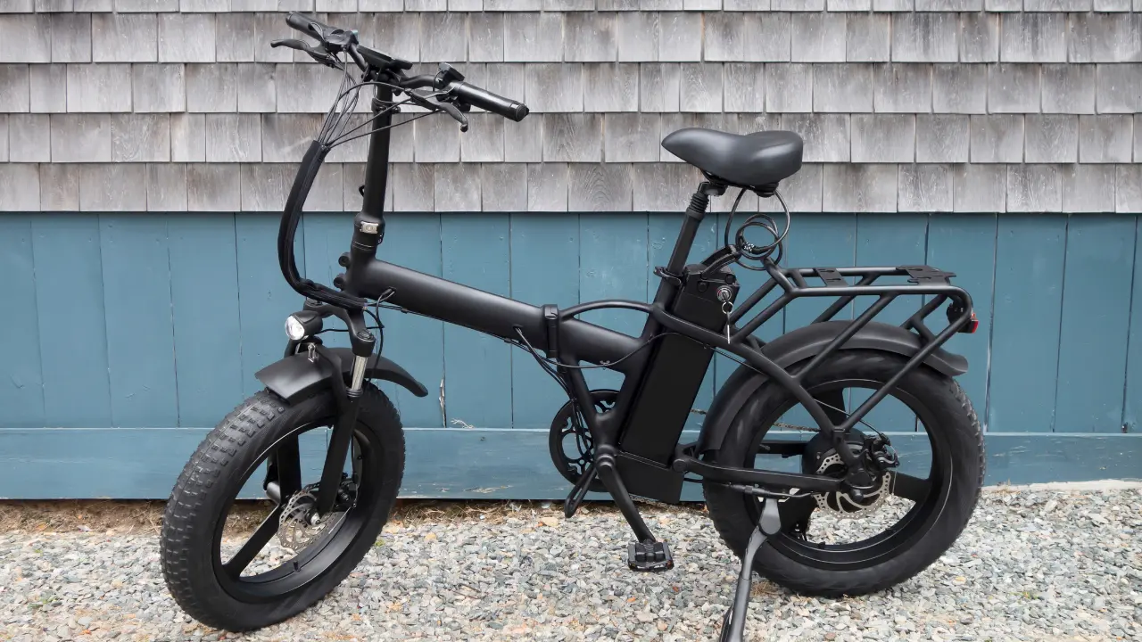 Imagem de uma bicicleta elétrica na cor preta, com pneus largos e espessos, equipada com um porta-bagagens na parte traseira e uma bateria visível montada no quadro. A bicicleta está estacionada em uma superfície de cascalho, com uma parede de madeira pintada ao fundo.