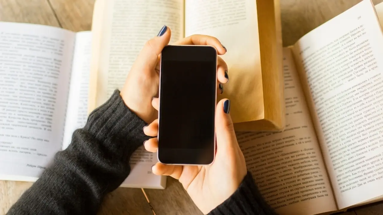 Imagem de uma pessoa segurando um smartphone com a tela apagada, posicionada entre as páginas abertas de dois livros grandes. As mãos, com unhas pintadas de escuro, aparecem sobre uma mesa de madeira clara. O foco está no celular, enquanto os livros, com texto legível, formam o pano de fundo, sugerindo uma interação entre a leitura física e digital.