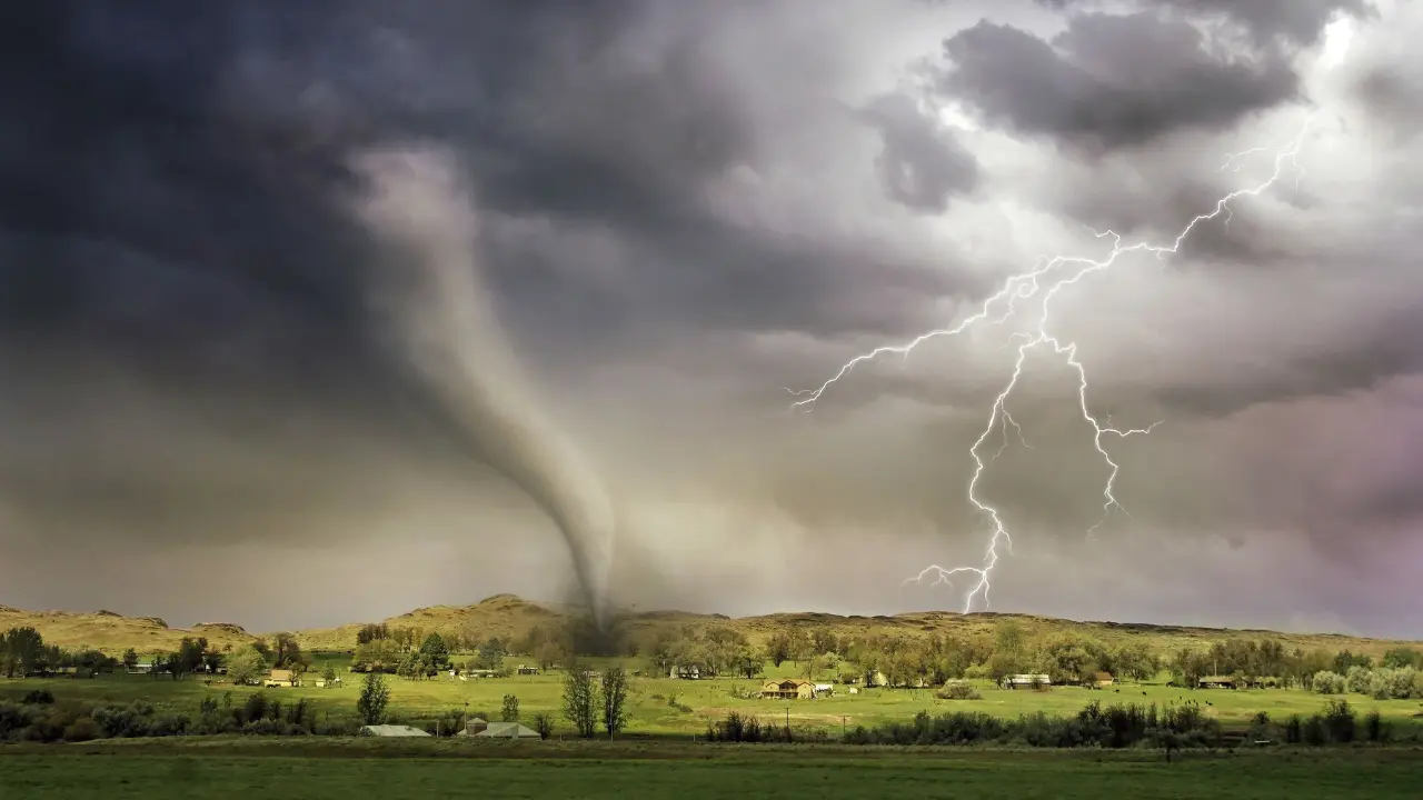 Uma paisagem rural sendo atingida por um tornado, com uma nuvem de tempestade escura no céu e um raio visível ao fundo. A imagem mostra o tornado tocando o chão próximo a uma pequena vila cercada por campos verdes.