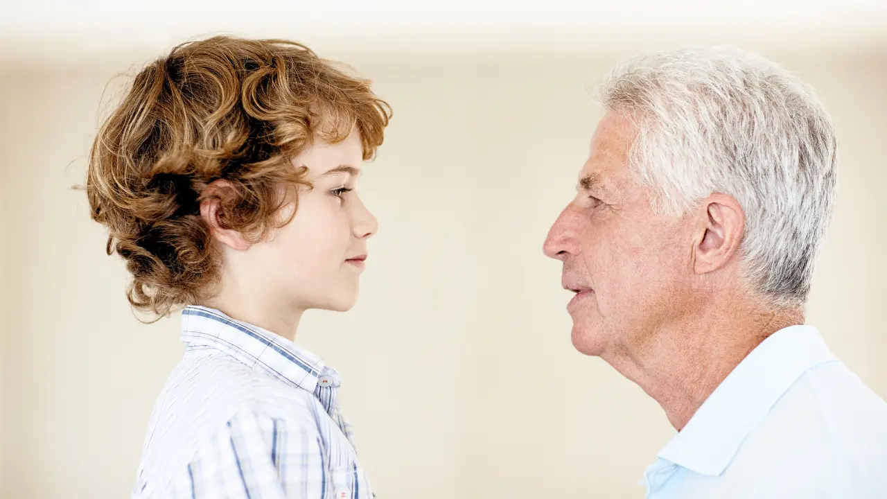 Um jovem menino de cabelos cacheados olha para um homem mais velho de cabelos grisalhos, ambos de perfil, frente a frente. O menino veste uma camisa xadrez e o homem uma camisa clara. A cena transmite um encontro de gerações, com ambos se encarando de maneira respeitosa e atenta.