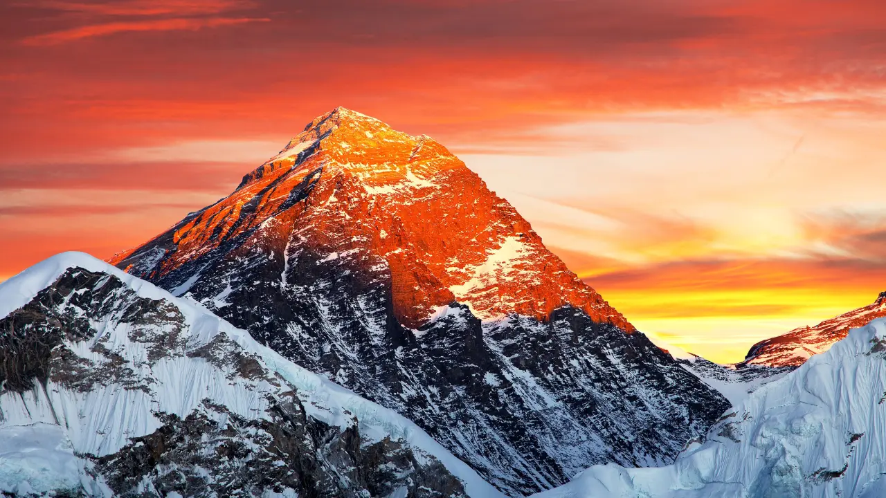 Imagem do Monte Everest ao pôr do sol, com o topo da montanha coberto de neve e iluminado por uma luz alaranjada intensa. A paisagem exibe o contraste entre as encostas escuras da montanha e o céu vibrante em tons de laranja e amarelo ao fundo.