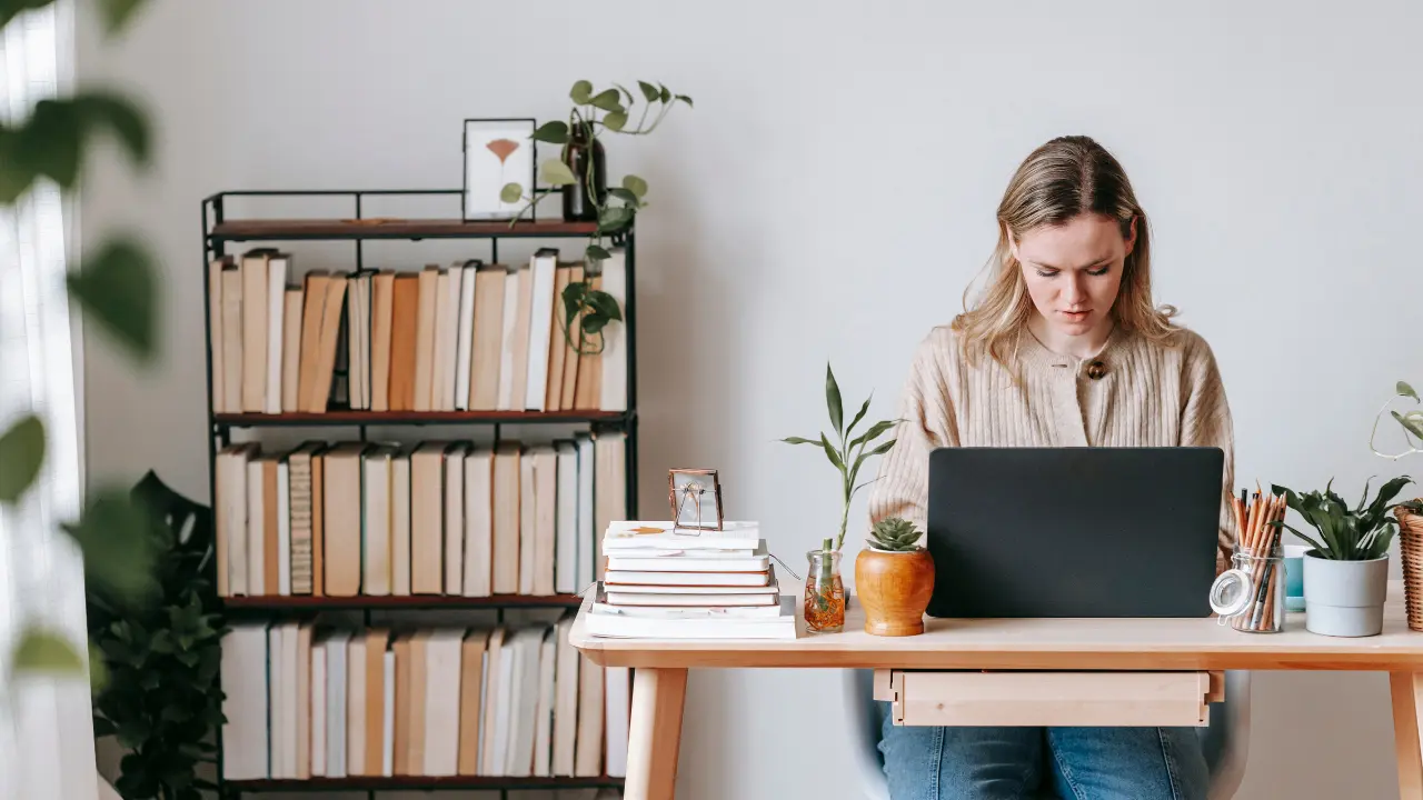 Mulher sentada à mesa de trabalho com um laptop, cercada por plantas e livros, em um ambiente de estudo com prateleira ao fundo repleta de livros.