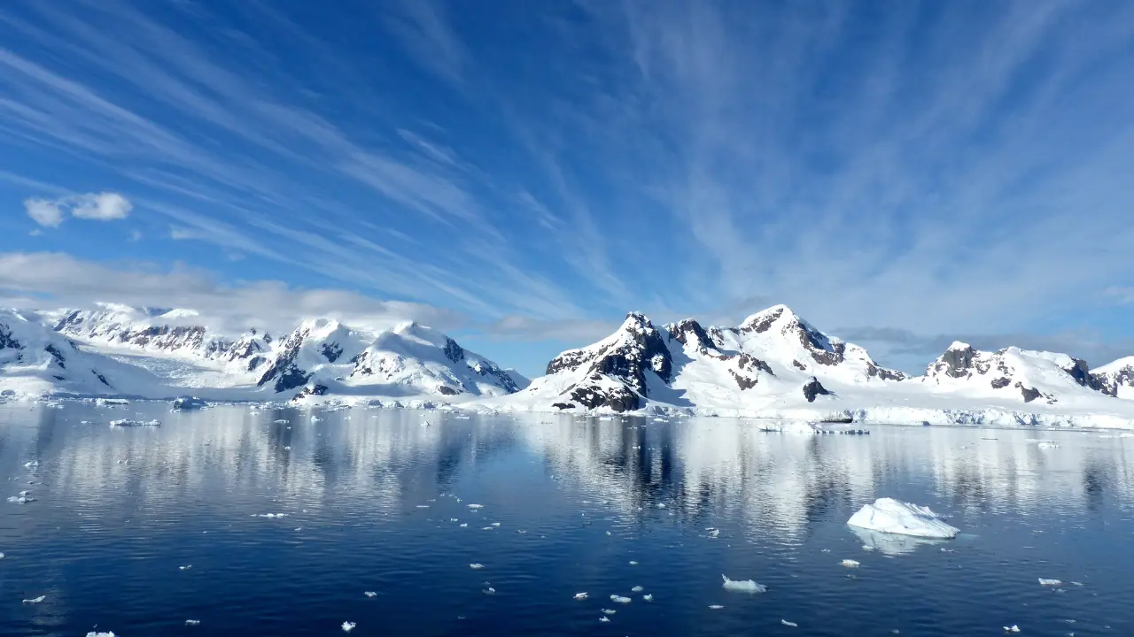 Imagem de uma paisagem da Antártida: montanhas cobertas de neve refletem na superfície tranquila e espelhada do mar, com blocos de gelo flutuando. O céu azul está adornado com nuvens finas e espalhadas.