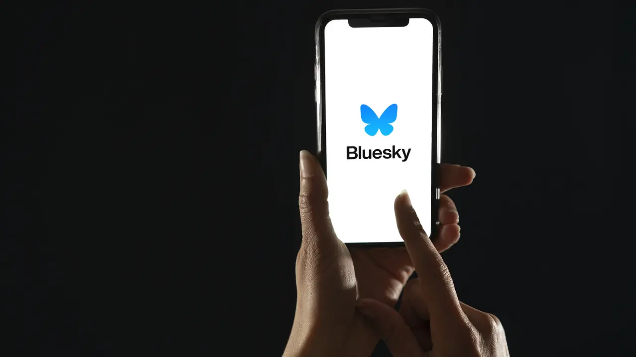 Mãos segurando um celular com a logo da Bluesky na tela. O fundo da imagem é preto.