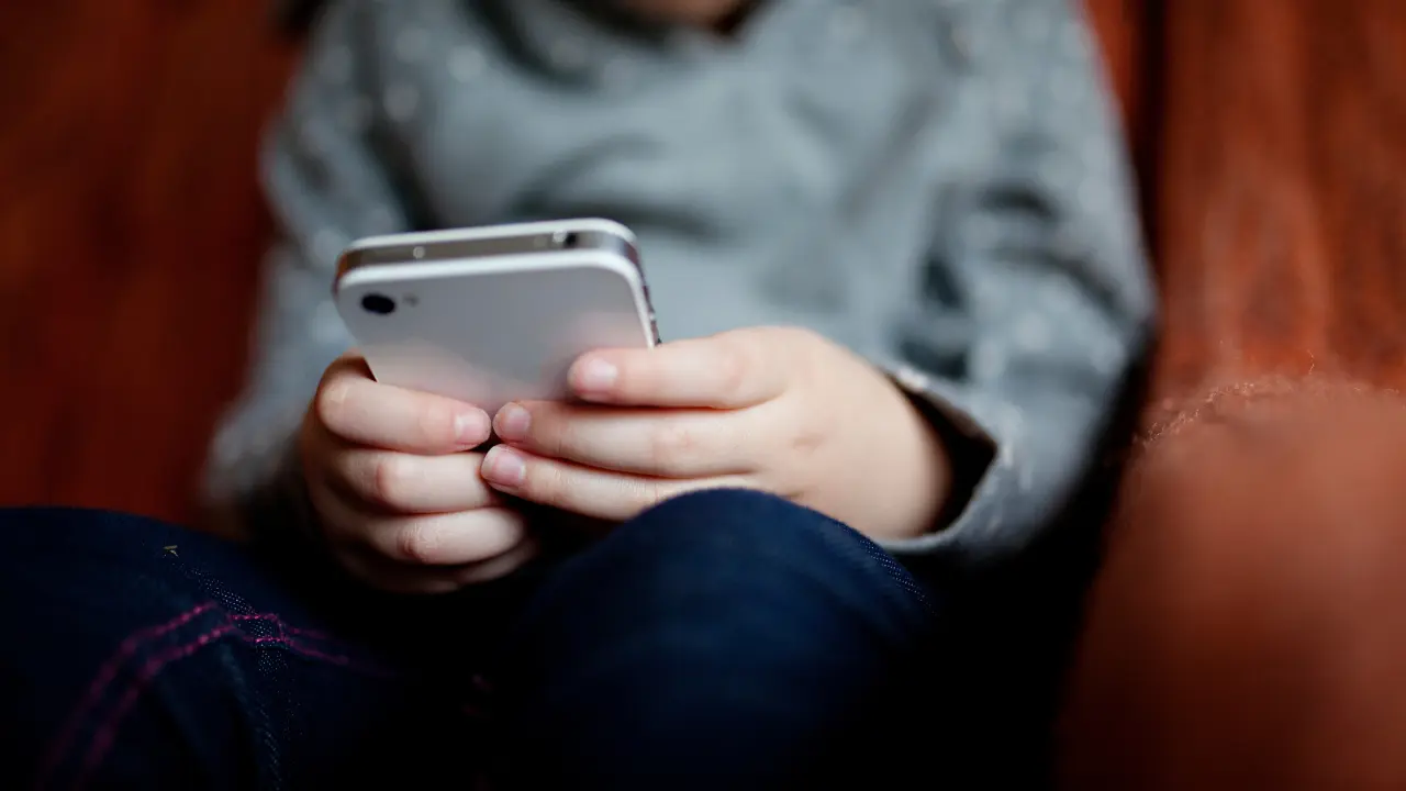 Uma criança segurando um celular com as duas mãos, concentrada na tela. Ela veste uma camisa cinza e está sentada, com foco nas mãos e no aparelho, enquanto o rosto não está visível.