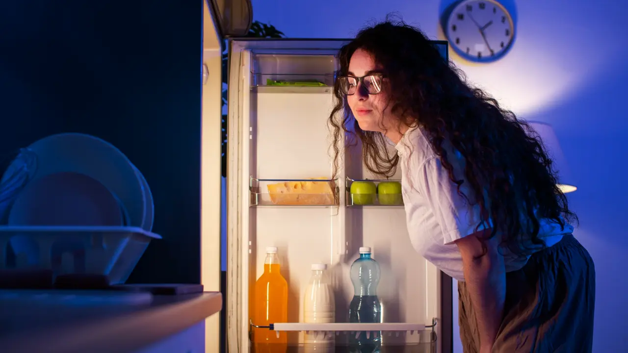 Mulher de cabelos longos e ondulados, usando óculos e camiseta branca, está olhando dentro de uma geladeira aberta à noite. A cena está iluminada pela luz da geladeira, e ao fundo há um relógio de parede marcando um horário noturno. Na geladeira, há garrafas e frutas dispostas nas prateleiras