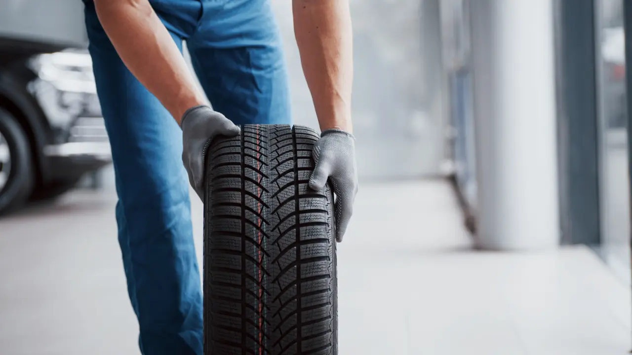 Um mecânico com luvas segurando um pneu novo em uma oficina automotiva, pronto para instalação. O pneu apresenta um padrão de ranhuras para aderência e estabilidade.